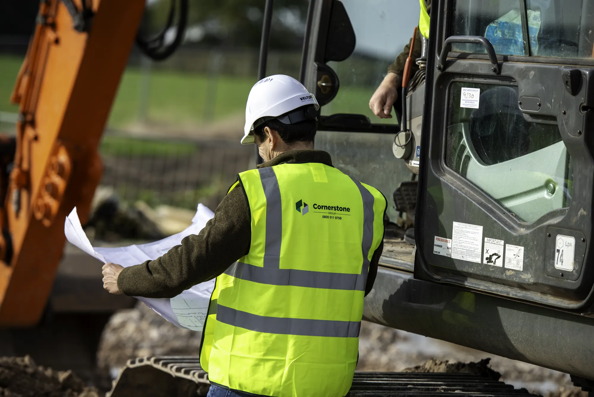 Cornerstone manager in hard hat and branded hi-vis checking plans beside an excavator