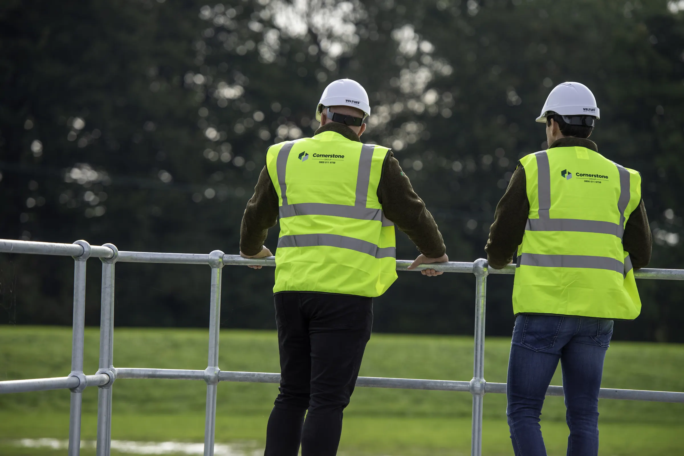 Two Cornerstone staff in hard hats and branded hi-vis at railings overlooking a utilities site
