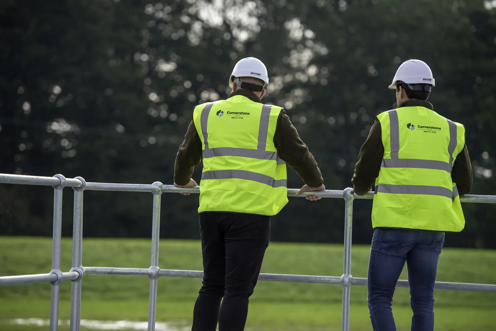 Two Cornerstone staff in hard hats and branded hi-vis at railings overlooking a utilities site